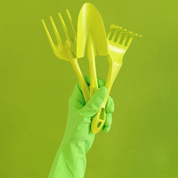Crop anonymous person wearing rubber glove showing set of gardening tools including trowel and hand rake against green background